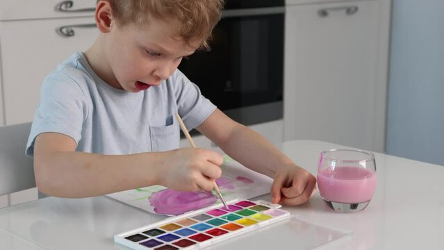 Blond haired cute boy paints using a brush and pink paint while sitting at a white table at home in kitchen. Boy learns to draw with watercolor paints. First steps of Caucasian boy in the visual arts