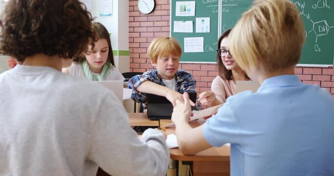 Caucasian cheerful kids sitting at desk with young female teacher and learning, doing tasks of homework or playing. School classes concept. Interctive studying. Education. Children talking with master