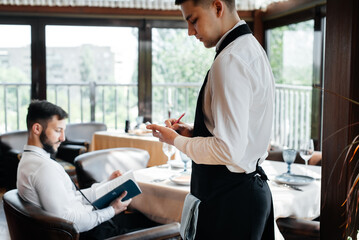 A young businessman in a fine restaurant examines the menu and makes an order to a young waiter in...