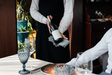 A young sommelier in a stylish uniform demonstrates and offers the client fine wine in the restaurant. Customer service. Table setting in a fine restaurant.