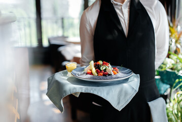 Close-up of a young waiter in a stylish uniform carrying an exquisite salad to a client in a beautiful gourmet restaurant. Table service in the restaurant.