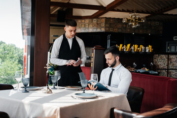 A young businessman in a fine restaurant examines the menu and makes an order to a young waiter in a stylish apron. Customer service. Table service in the restaurant.