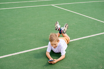 The boy is lying on the green grass for football and playing on the phone
