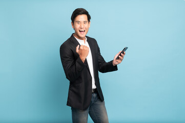 Young asian man in suit and jeans using smartphone exciteing with news on phone for smart casual business concept isolated on blue background