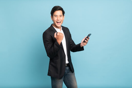 Young Asian Man In Suit And Jeans Using Smartphone Exciteing With News On Phone For Smart Casual Business Concept Isolated On Blue Background