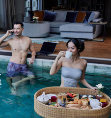 Happy Asian couple having breakfast in the pool. Floating breakfast tray in luxury hotel. Young...