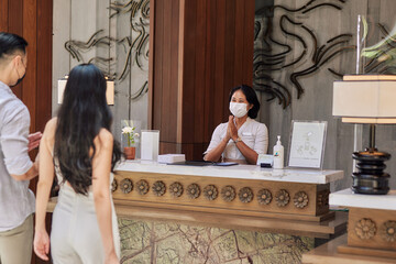 Young couple wearing medical masks as precaution against virus, walking in lobby and arriving at hotel reception while front desk employee welcoming and greeting them - obrazy, fototapety, plakaty