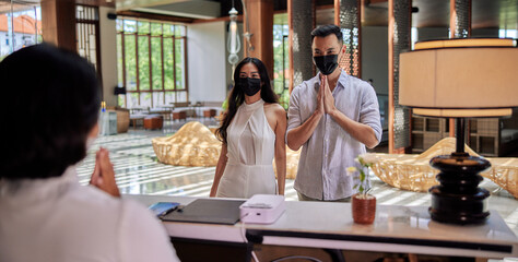 Young couple wearing medical masks as precaution against virus, walking in lobby and arriving at hotel reception while front desk employee welcoming and greeting them