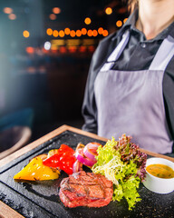 Waiter in an apron carries an order on a dish in a restaurant. Served grilled steak with vegetables on the board. Service in the restaurant