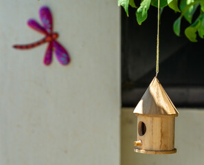Wooden birdhouse hanging from a tree branch in the garden with the colorful silhouette of a decorative dragonfly on the wall