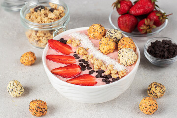 Smoothie bowl with strawberries, granola, chocolate, coconut flakes and crunchy honey balls in white bowl on gray background