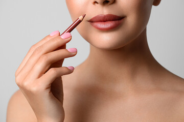 Young woman applying beautiful nude lip pencil on light grey background, closeup