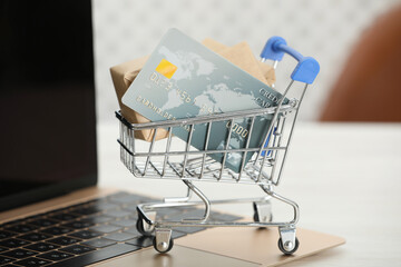 Online payment concept. Small shopping cart with bank card, boxes and laptop on white table, closeup