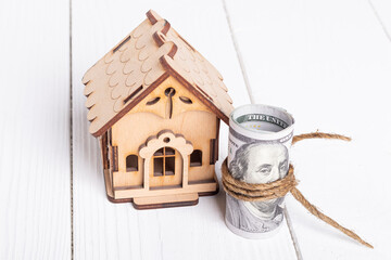 white wooden background in retro style. a toy house and dollars wrapped in a roll next to it. a thread from a drape.