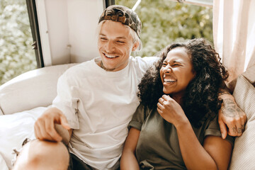 Mixed-race couple chilling on white sofa, laughing and hugging, enjoying their relationship's vibes, African woman feeling happy sitting next to her Caucasian hipster man, both dressed casually