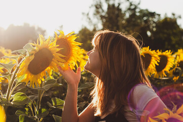a beautiful young girl with them red hair smells a flower in a field of sunflowers at sunset,...