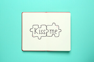 Open notebook with phrase Kiss Me and drawn puzzles on turquoise background, top view
