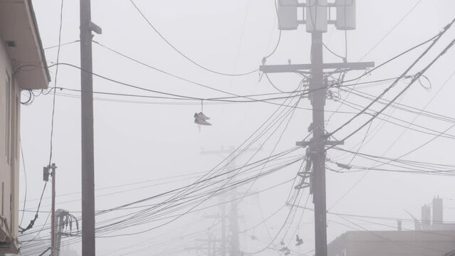 Sneakers canvas shoes hanging on power line, fog on city street, misty foggy weather in California, USA. Pair of boots dangling on laces or shoelace on cable or wire. Gloomy moody dramatic atmosphere.