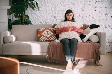 Obraz premium A young woman sits comfortably on a sofa in a modern bright apartment. A beautiful brunette girl in a knitted sweater is relaxing at home