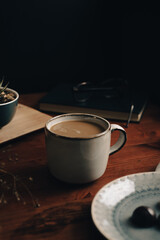 Taza de café con bombones de chocolates en un plato, con libro verde con gafas, lentes encima, planta, mesa de madera, paño blanco, en fondo oscuro