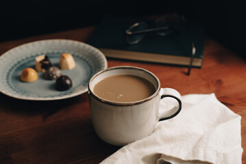 Taza de café con bombones de chocolates en un plato, con libro verde con gafas, lentes encima, mesa de madera, paño blanco, en fondo oscuro