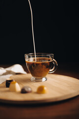 Taza de café, taza de vidrio sirviendo leche, en mesa de madera, encima de bandeja de madera con bombones de chocolate, mientras cae leche