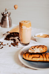 Vaso de Café Dalgona, Delicioso café coreano, café frío, con fondo blanco, rodeado de granos de café, bollos dulces de chocolate, y molino de café plateado