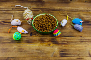 Set of toys for cat and bowl with dry pet food on wooden background. Pet care concept