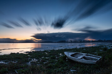 Long exposure view of shore of Trasimeno lake with a little boat at dusk, with perfectly still water and moving clouds