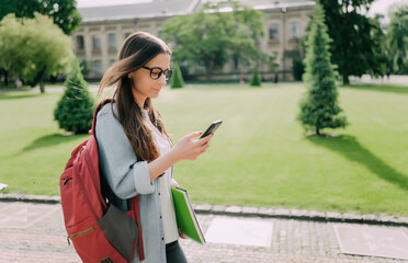 Fototapeta premium Cute student girl walking and loock at mobile phone with a university building in the background