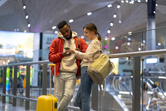 Young european woman with smartphone showing direction to african man traveler in airport. Couple of friends travelers looking at mobile phone screen while traveling together, searching location - Powered by Adobe