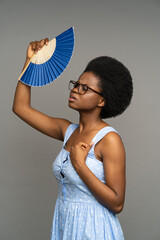 Fototapeta Young black woman suffering from heat or high air temperature inside, waving with paper fan standing isolated on grey. Overheated african american female fanning herself to cool down on hot summer day obraz