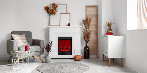 Interior of light room with modern fireplace, chest of drawers, armchair and autumn decor