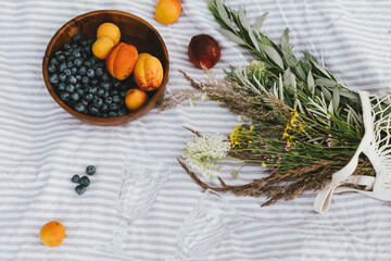 Summer picnic and vacation concept. Tasty fruits and berries, wineglasses and bouquet of wildflowers on blanket, top view. Blueberries, peaches and apricots