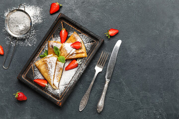 Board with thin pancakes, strawberries and sugar powder on dark background