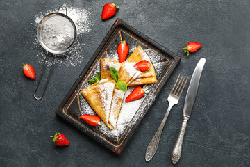 Board with thin pancakes, strawberries and sugar powder on dark background