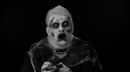 Sinister man with horrible scary Halloween zombie makeup in convulsions making faces, looking ominous, trying to scare. Dead guy with wounded bloody scars face isolated against black wall background