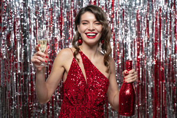 Happy party woman in red dress holding bottle and glass of champagne on tinsel background