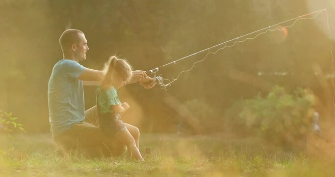 Father throws fishing rod into river to catch fish and daughter helps in park. Dad with girl likes fishing together at blurry sunset slow motion