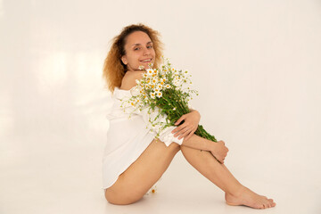 Young slim happy caucasian woman sits with bare legs on the floor. Bouquet of field daisies in hands. No makeup. Naturalness, body positivity, freedom. Matricaria recutita. Mental health care. 