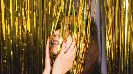 Young woman hiding face under golden tinsel in studio. Portrait of female with bright manicure on...
