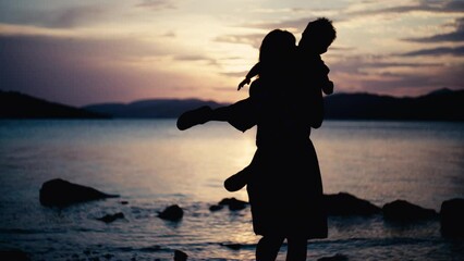 A slow motion video of a mother spinning her son in her arms by the sea at sunset. Happy family having fun together on the ocean beach on vacation.