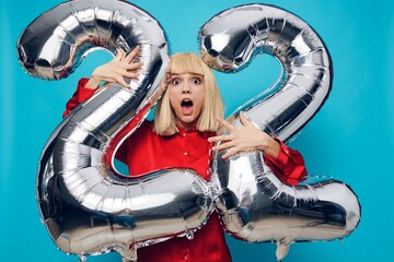 a bright, emotional, funny woman in a red shirt stands on a blue background and holds inflatable balloons in the shape of the number twenty-two in silver color, shouting loudly with delight