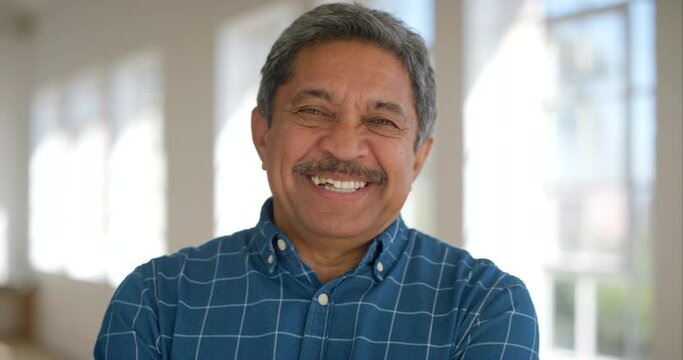 Portrait of a happy retired male looking cheerful standing inside a bright room. Face of a laughing mature latino man standing in an empty office or home. Smiling confident senior enjoying retirement
