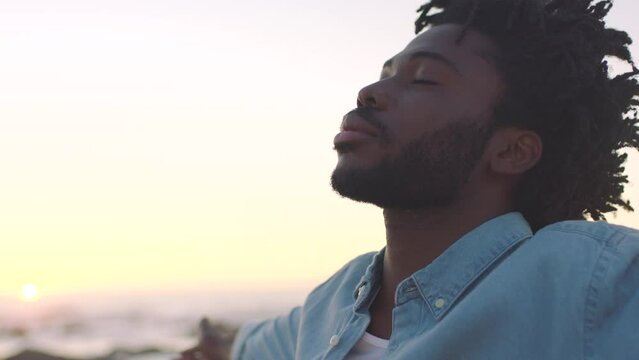 Closeup of an African American man finding inner peace at sunset, tranquil and quiet at a beach with copyspace. Black male meditating and enjoying harmony with nature, zen and calm breathing exercise