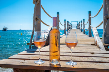 Obraz premium Glasses and bottle of cold rose wine from Provence served outdoor on wooden yacht pier with view on blue water and white sandy beach Plage de Pampelonne near Saint-Tropez, France