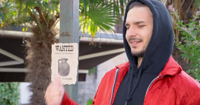 Suspicious-looking young man in hood sneers contemptuously, pointing his finger at ransom notice for antique jug hanging on pole, front view. Guy is stirring up something illegal