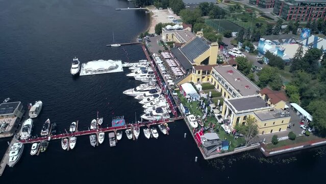 Exciting aerial view of marina with moored yachts and pier with walking people on it. The high view of the trees, tennis court and city infrastructure.