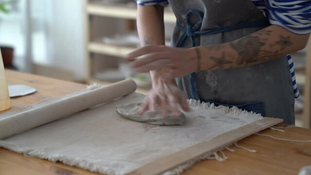 Master pottery wearing apron wedging clay with hands without using electric potters wheel, standing behind table in studio. Process of making pottery, close up. Hand-kneading technique in ceramics
