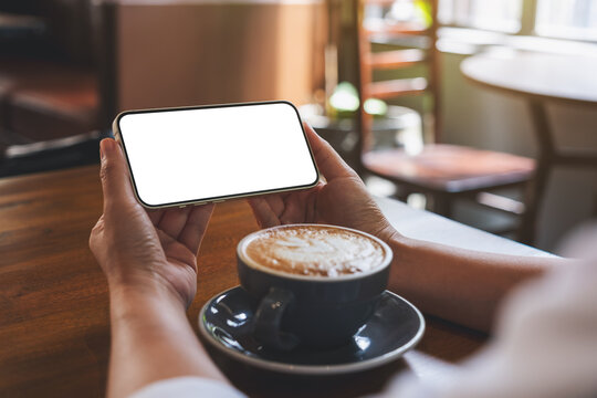 Mockup image of hands holding mobile phone with blank desktop screen with coffee cup on wooden table